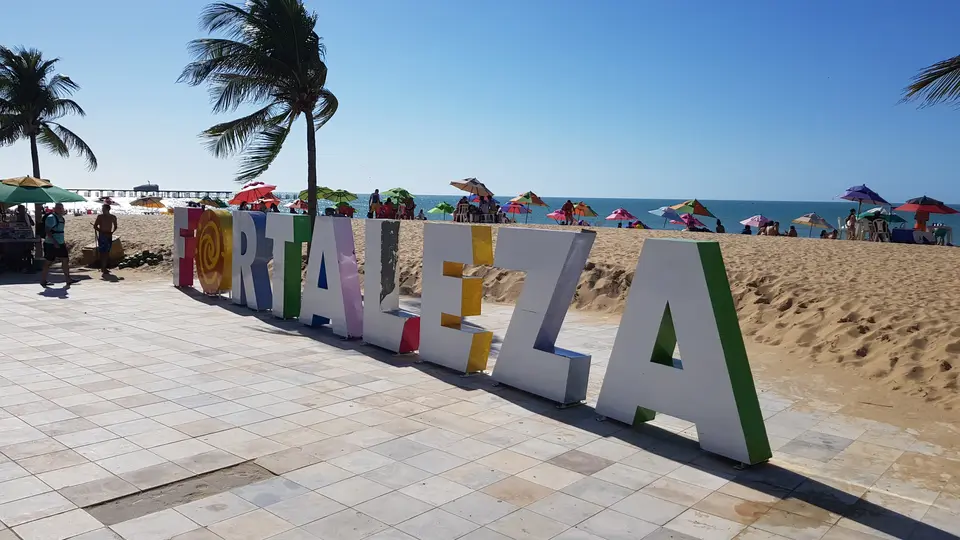 Bunte FORTALEZA-Buchstaben am Strand mit Palmen und Sonnenschirmen im Hintergrund